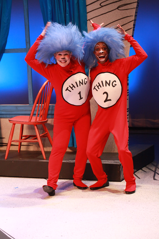 Two actors raise their fists in the air on stage while costumes in blue wigs and red onesies that read 