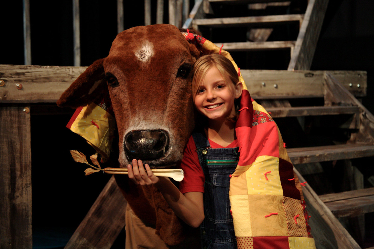 A youth actor holds a celery prop up to a large cow puppet face while both she and the cow have a quilt draped over them.