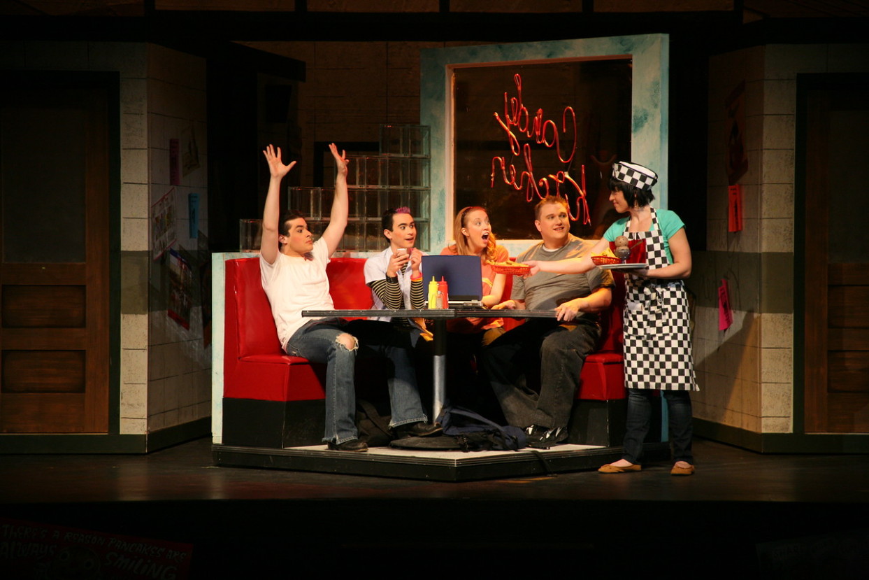 Four actors sit in a diner booth set on stage while a waitress in a checkered apron serves them.