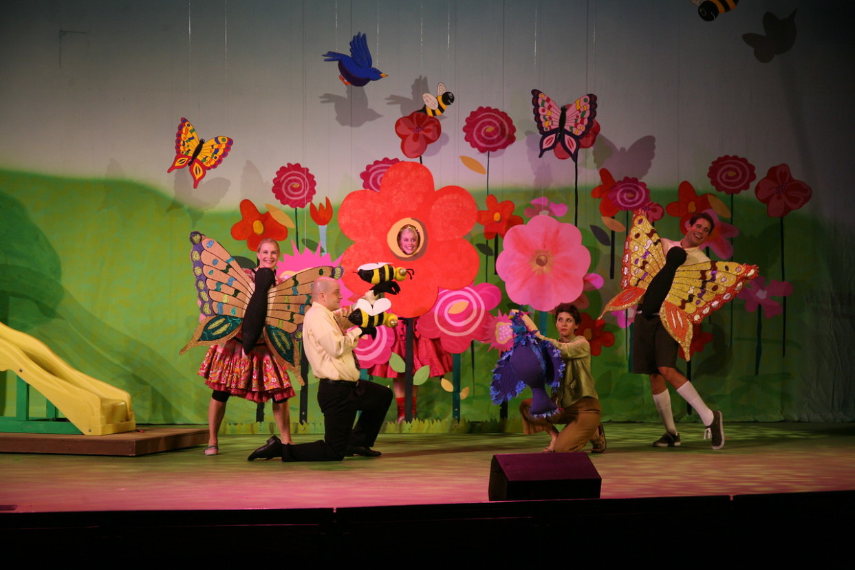Four actors hold butterfly, bumblebee and bird puppets in front of a bright oversized flower garden set piece on stage.