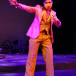 Photo description: A male actor wears a sparkly pink blazer over a gold vest and pants with a pink button-up shirt. His left arm is outstretched and his right arm is bent near his chest in a dance move.