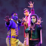 Photo description: Three actors strike a dramatic pose with their arms outstretched. A male actor is wearing a blue military-inspired jacket over a yellow and blue jumpsuit with bright blue boots and a blue mask. A female actor is wearing a greet jacket embellished with pink sequins and long pink fringe along the sleeves and back of the jacket. She wears striped grey pants with pink sequin trim and pink high-heeled boots. Another male actor wears a sparkly pink blazer over a gold vest and pants with a pink button-up shirt.