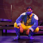 Photo description: A male actor wearing a blue military-inspired jacket over a yellow and blue jumpsuit with bright blue boots and a blue mask crouches down near a set of stairs while looking at the camera.