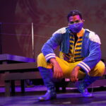 Photo description: A male actor wearing a blue military-inspired jacket over a yellow and blue jumpsuit with bright blue boots and a blue mask crouches down near a set of stairs while looking at the camera.